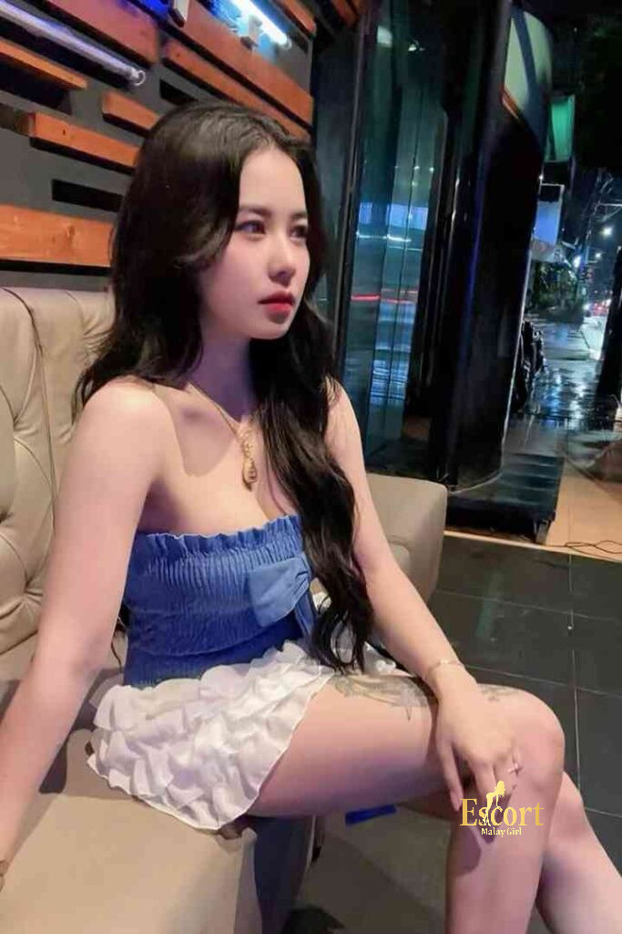 Escort Malay Girl Adeline, a 22yo C Cup Thailand Escort in KL, sitting on a couch wearing a blue top and a white skirt, with an "Escort Malaysia" watermark KL Escort Girl Service
