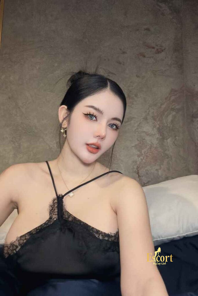 Escort Malay Girl Grace Thailand Escort in black lace dress, sitting on a bed with pillows KL Escort Girl Service