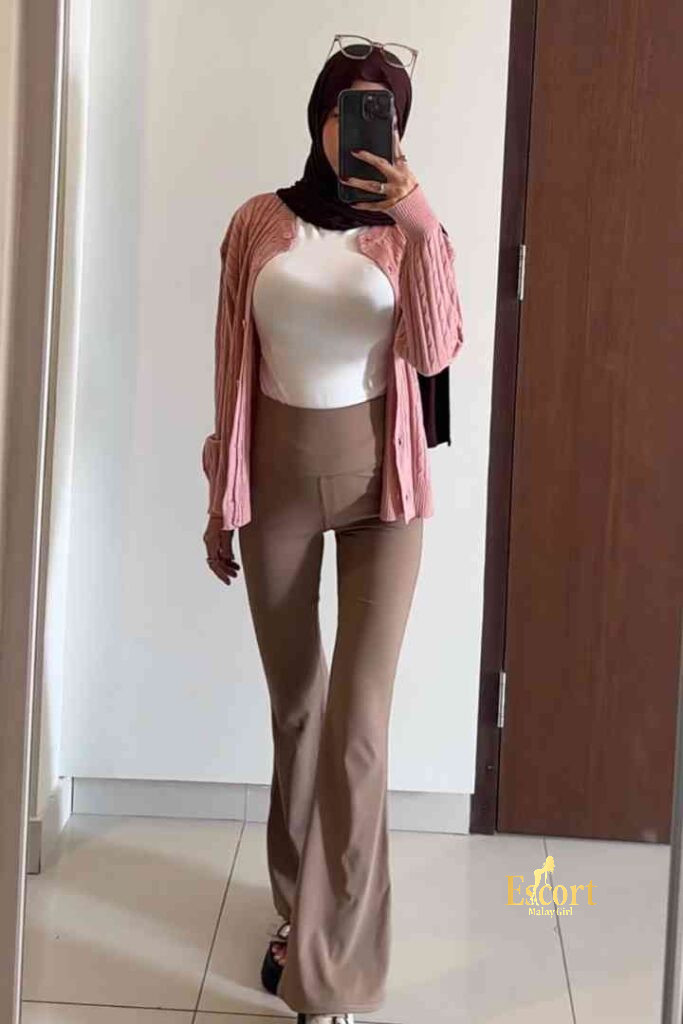 Escort Malay Girl Kirana, a Malay escort in KL, takes a selfie wearing a pink cardigan and brown pants. KL Escort Girl Service