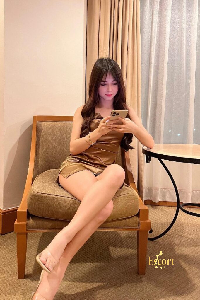 Escort Malay Girl Zanis, 22yo Malay escort in a brown dress, sitting on a chair looking at her phone KL Escort Girl Service